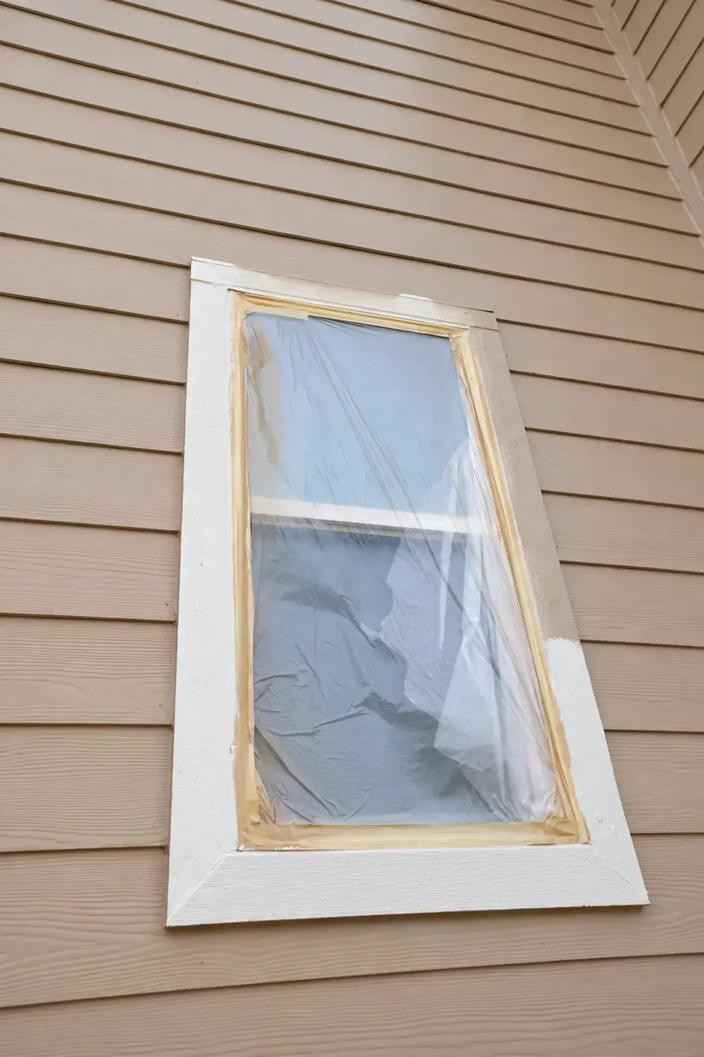 A separate exterior window with its glass fully covered by plastic during painting preparation
